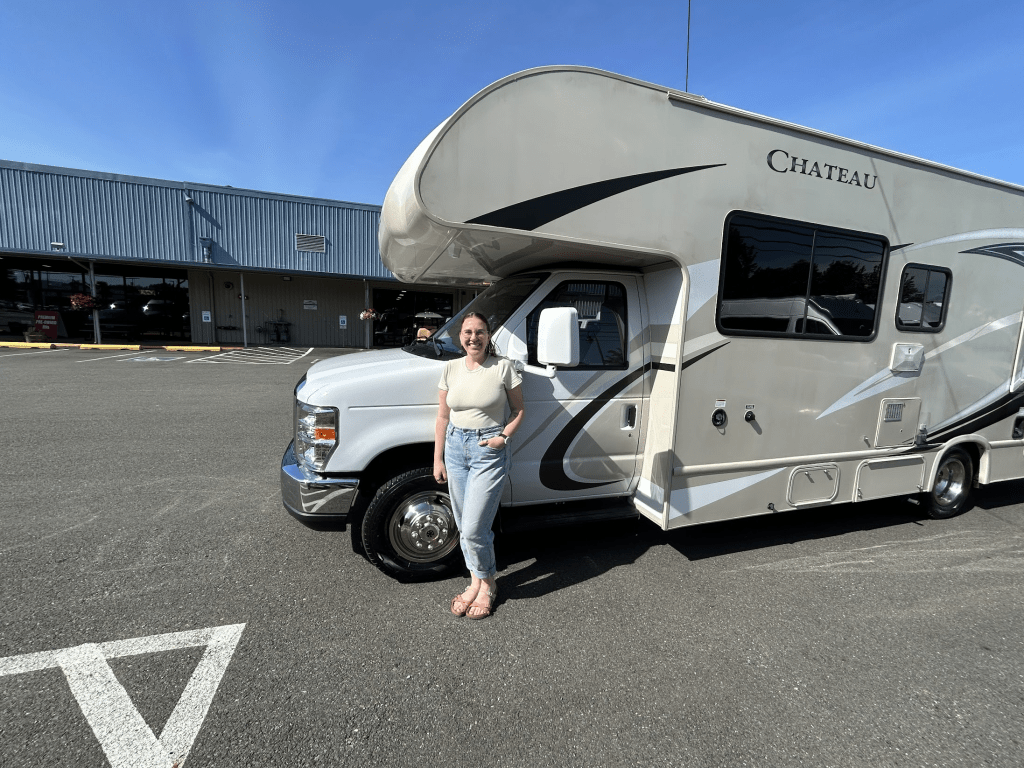 Hannah stands happily next to her beige recreational vehicle (RV) labeled 'Chateau' in a parking lot, with the dealer's building and blue sky in the background.